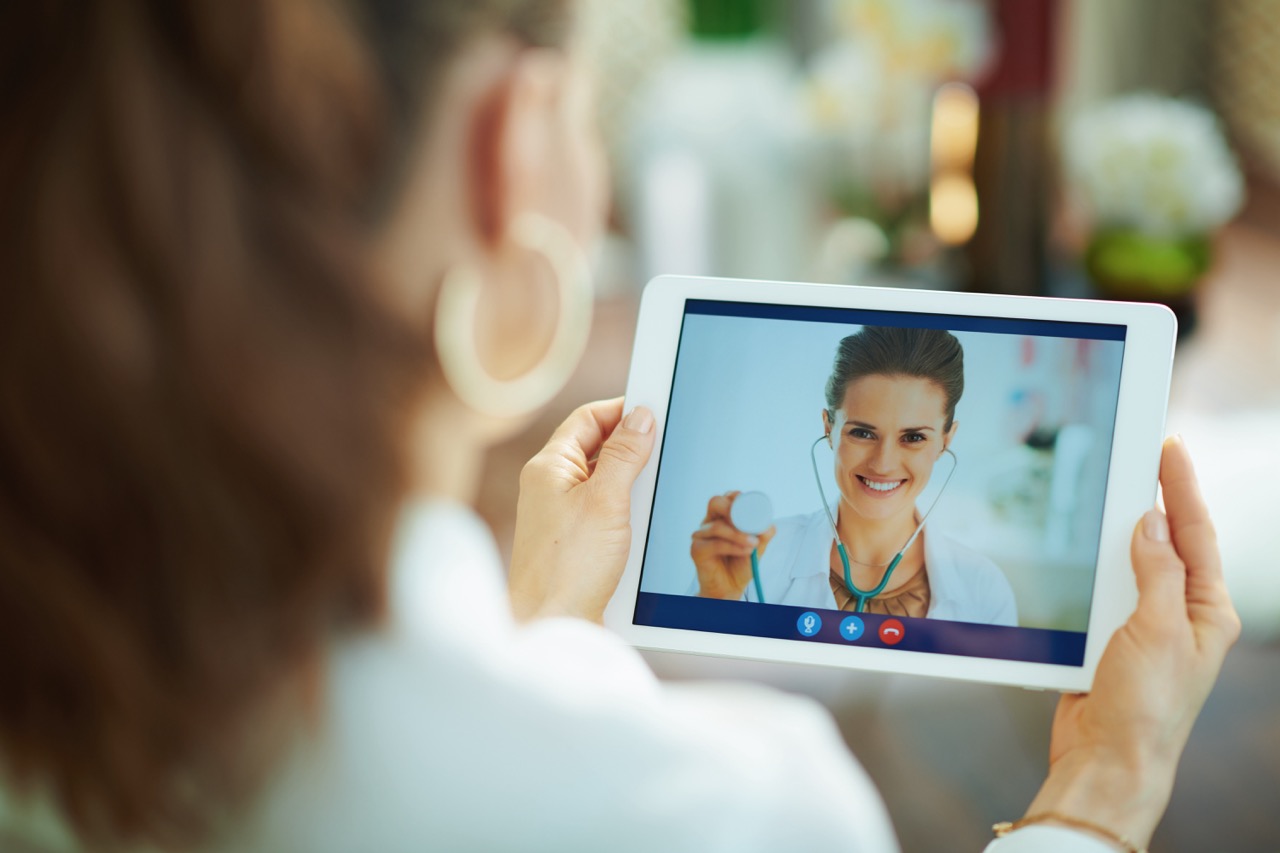 Seen from behind female speaking with doctor using telehealth technology on a tablet PC in the living room in sunny day.